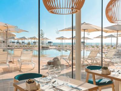 Großes Fenster mit Blick auf Pool und Sonnenliegen, helle Holzmöbel, weiße Sonnenschirme draußen.