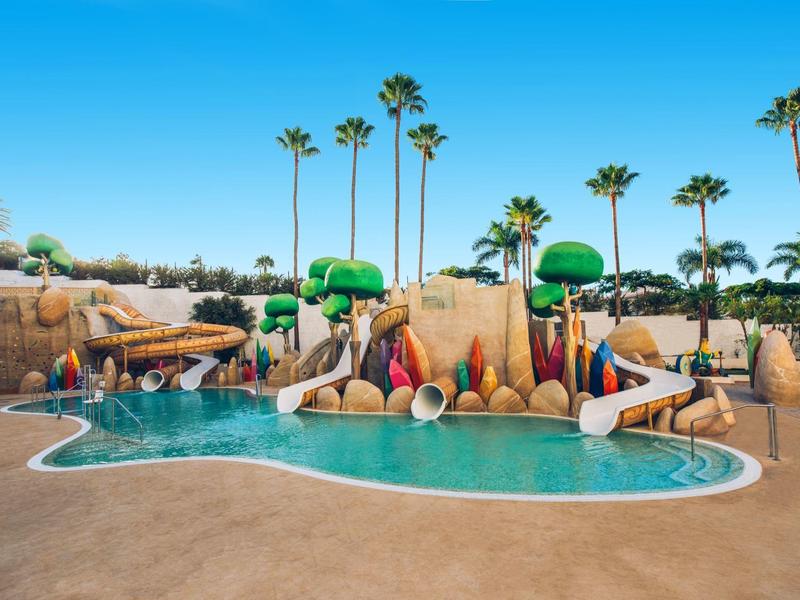 Parque acuático con toboganes y palmeras junto a una piscina en un complejo vacacional.