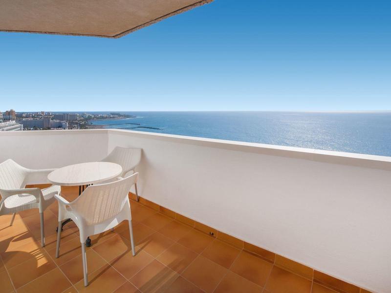 Balcón con sillas blancas y mesa redonda, vista al mar bajo cielo despejado.