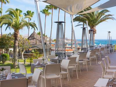 Terrasse mit Tischen und Stühlen, Palmen und Blick aufs Meer im Hintergrund.