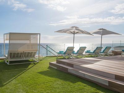 Lounge chairs and umbrellas by a pool overlooking the sea under a partly cloudy sky.
