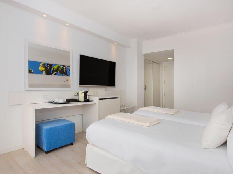 Bright hotel room with two beds, TV, desk, and blue stool.