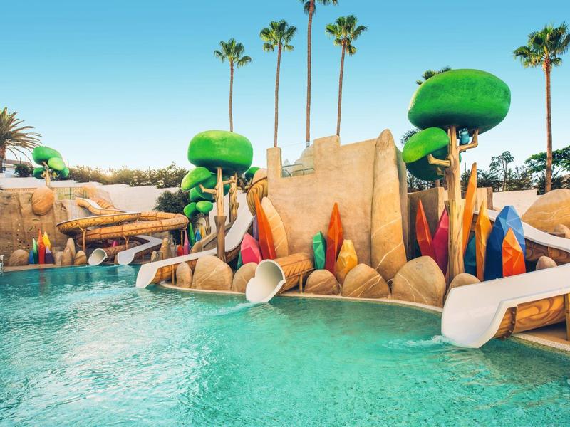 Colorful water playground with slides and artificial trees surrounded by clear water.
