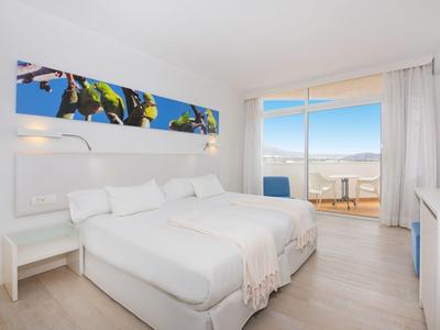 Lichte hotelkamer met twee bedden en balkon met uitzicht op zee onder een zonnige lucht.