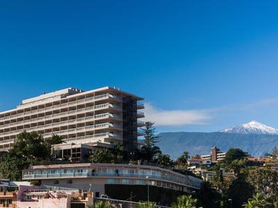 Bâtiment d'hôtel moderne avec balcons sous un ciel bleu et une montagne enneigée en arrière-plan.