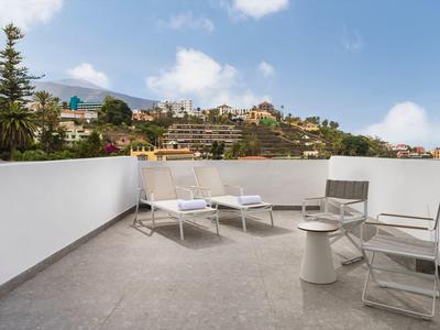 Balcone con sedie e lettini che si affaccia su case e colline verdi in una giornata di sole.