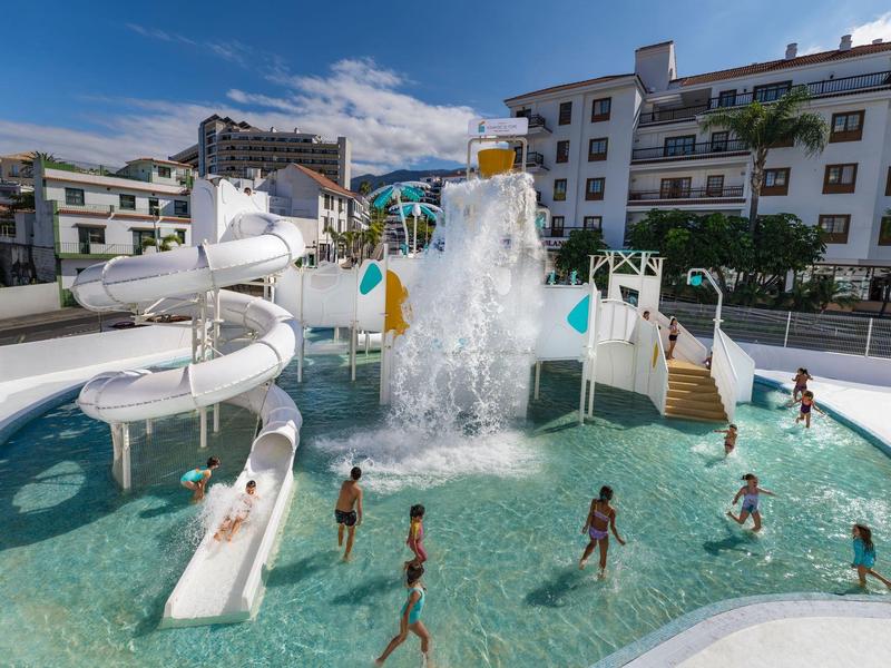 Des enfants jouent et glissent dans une piscine extérieure d'hôtel avec toboggans et une cascade d'eau.