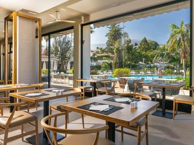 Bright restaurant with wooden tables overlooking a pool and palm trees.