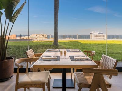 Dining area with table and chairs overlooking lawn and sea through large glass windows.