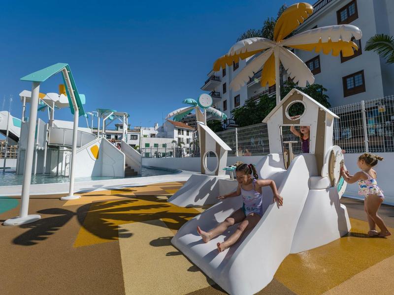 Children playing on a white slide in a sunny outdoor playground by a hotel.