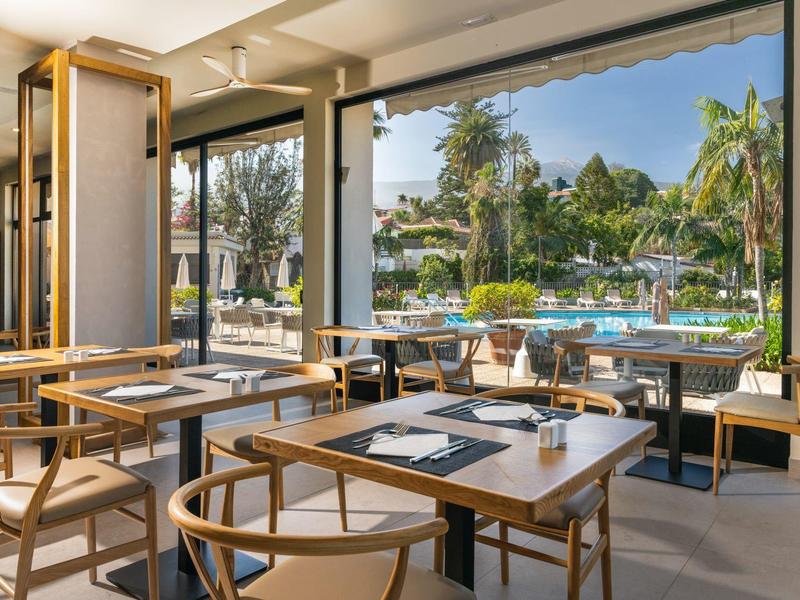 Bright restaurant with wooden tables overlooking a pool and palm trees.