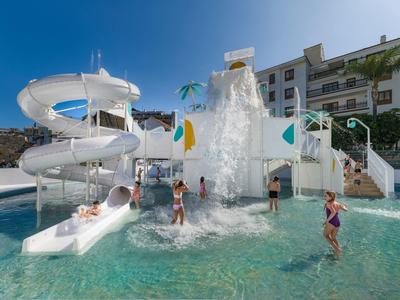Niños juegan en una piscina con tobogán de agua y cascada en un complejo hotelero bajo el sol.