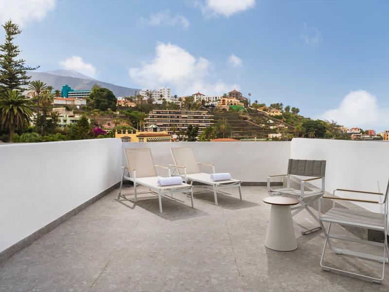 Balcone con sedie e lettini che si affaccia su case e colline verdi in una giornata di sole.