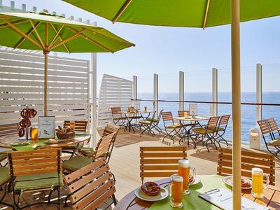 Sunny outdoor terrace with wooden tables, chairs, umbrellas, and sea view on a clear day.