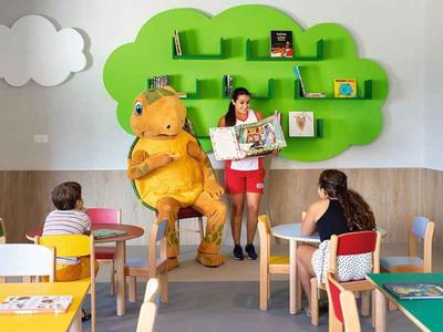 Sala de niños con mesas coloridas, estantería en forma de árbol y persona disfrazada de dinosaurio.