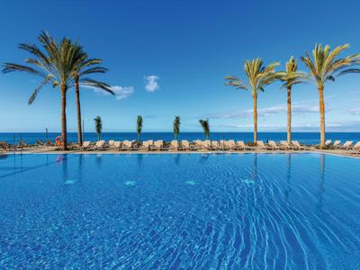 Piscina infinita azul con palmeras y vista al mar bajo un cielo despejado.