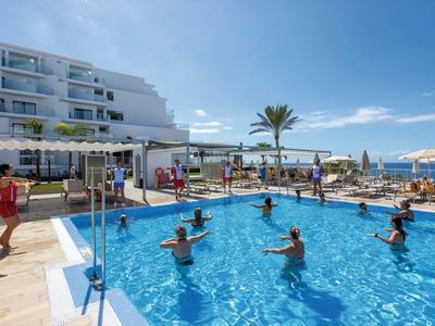 Grupo de personas haciendo aeróbicos acuáticos en una piscina de hotel cerca de la playa bajo un cielo azul claro.