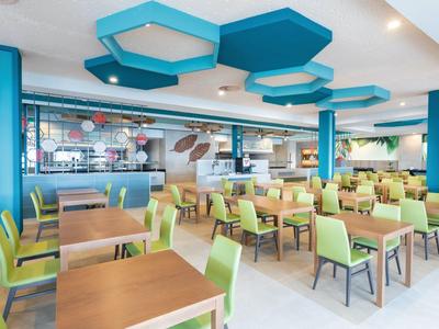 Lichte en moderne eethoek in hotel met groene stoelen, houten tafels en blauwe geometrische plafondpanelen.