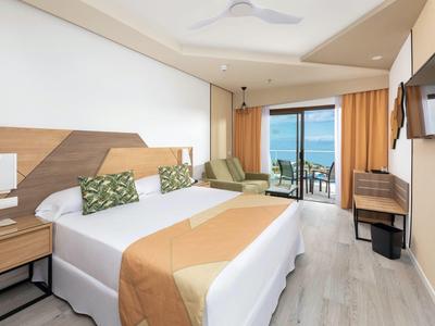 Helles Hotelzimmer mit Doppelbett, Holzdekor, grün gemusterten Kissen und Balkon mit Meerblick.