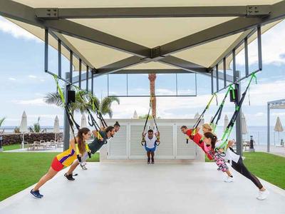 Mensen trainen met TRX-banden onder een overdekte buitenruimte aan zee.