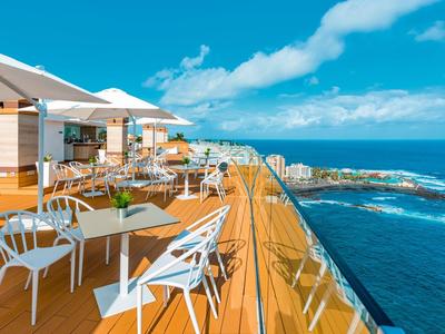 Terraza en la azotea con sillas y mesas blancas con vista al mar bajo un cielo soleado.