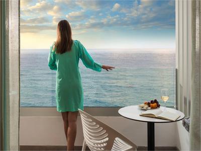 Vrouw in groene badjas kijkt vanaf balkon met tafel en snacks naar de zee.