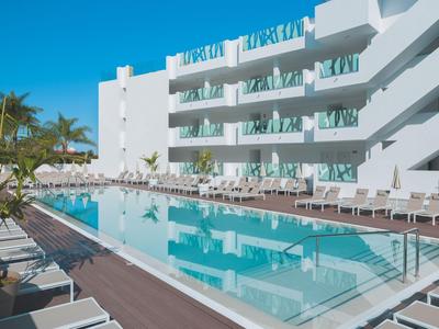 Modern hotelzwembad met ligbedden en witte parasols naast een wit gebouw onder een heldere lucht.
