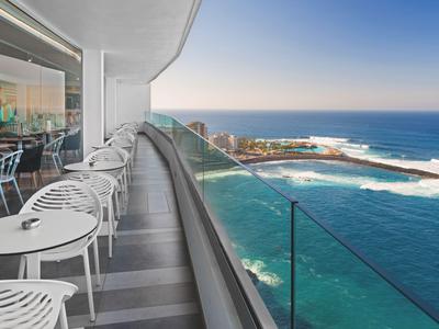 Balcon avec chaises et tables blanches donnant sur la mer et la côte au coucher du soleil.
