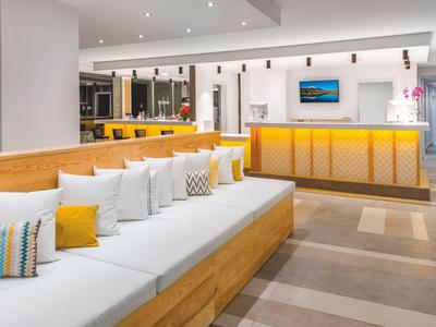 Hall d'hôtel moderne avec un long canapé à base en bois, des coussins blancs, des accents jaunes, comptoir de réception en arrière-plan.