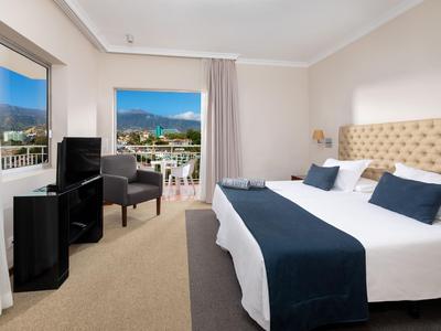 Chambre d'hôtel lumineuse avec lit double, coin salon et balcon avec vue sur la montagne.