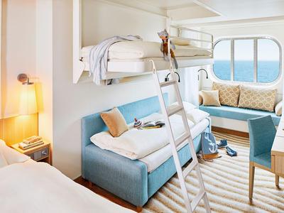 Bright cabin with bunk bed, sofa, seating area, and sea view through a round window.