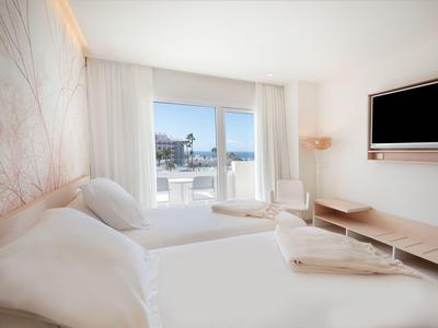 Modern hotel room with two beds, balcony, and sea view in daylight.