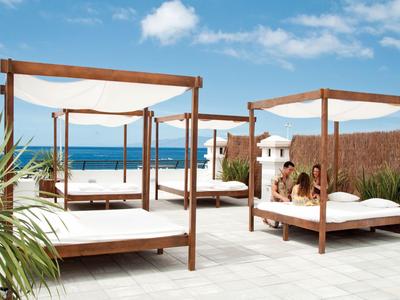 Letti lounge all'aperto moderni con tendaggi bianchi sotto un cielo azzurro vicino al mare.