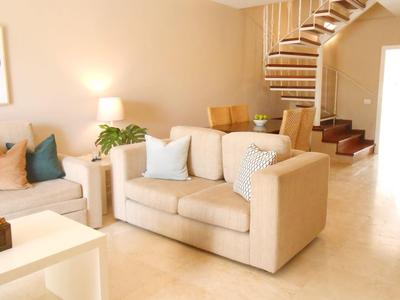 Bright living room with beige sofa, staircase, and small seating area in the background.