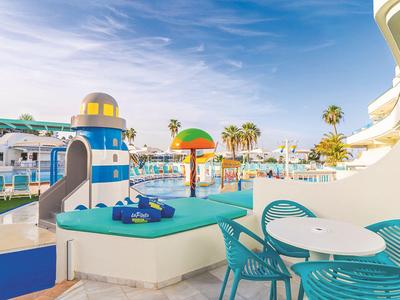 Blick auf eine sonnige Hotelterrasse mit Poolspielplatz und Meer im Hintergrund