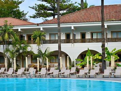 Hotel s bazénem v popředí, lehátka a palmy před dvoupodlažní budovou