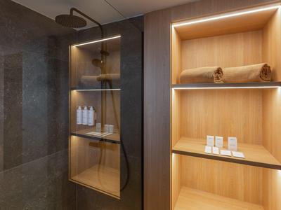 Bagno moderno con mensole in legno illuminate e soffione a pioggia in ambiente alberghiero.