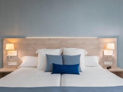 Letto matrimoniale con testiera in legno chiaro, cuscini bianchi e blu, lampade da parete, comodini.