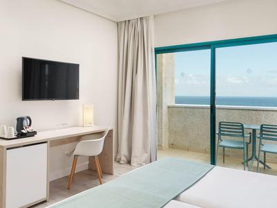 Habitación de hotel luminosa con cama, escritorio, televisor y balcón con vista al mar.