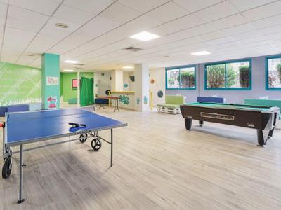 Sala de recreo luminosa con mesas de ping-pong y billar, grandes ventanas y suelo de madera.