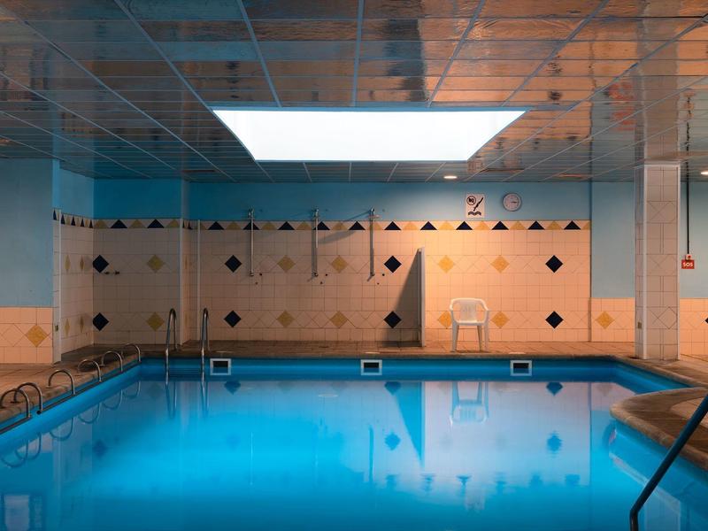 Piscina interior con agua iluminada en azul, pared de azulejos beige y techo espejado.