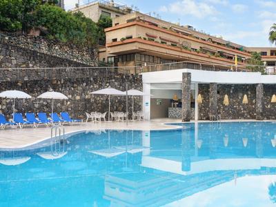 Piscine avec chaises longues bleues et parasols blancs à côté d'un hôtel à plusieurs étages.