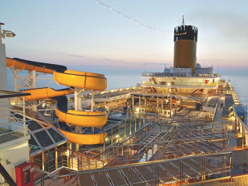 Large cruise ship deck at sunset with yellow water slides and open lounging areas.