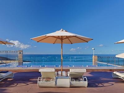 Pool area with sun loungers and umbrellas next to the sea under a clear sky.
