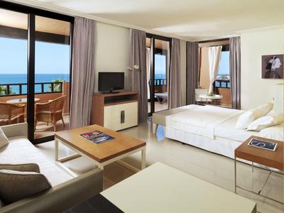 Bright hotel room with double bed, seating area, TV, and balcony with sea view.