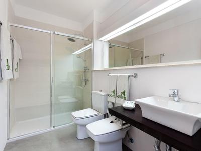 Baño moderno con ducha de vidrio, lavabo rectangular y bidé en tonos blancos y grises.
