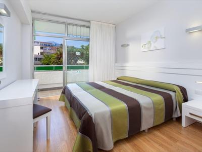 Habitación de hotel luminosa con cama doble, suelo de madera y balcón con vista verde.