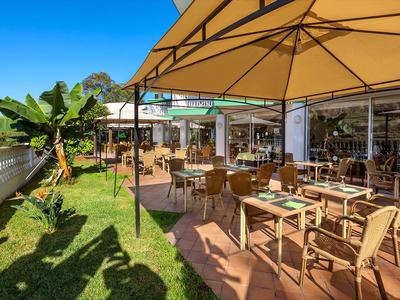 Terrasse ensoleillée avec tables et chaises à côté d'un restaurant et d'un jardin.