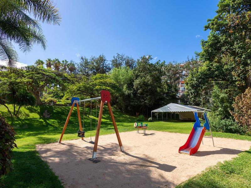 Terrain de jeu pour enfants avec balançoire et toboggan sur sable entouré de verdure sous un ciel bleu.