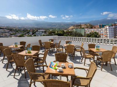 Rooftop terrace with wicker chairs and tables overlooking a city and mountains under a clear sky.